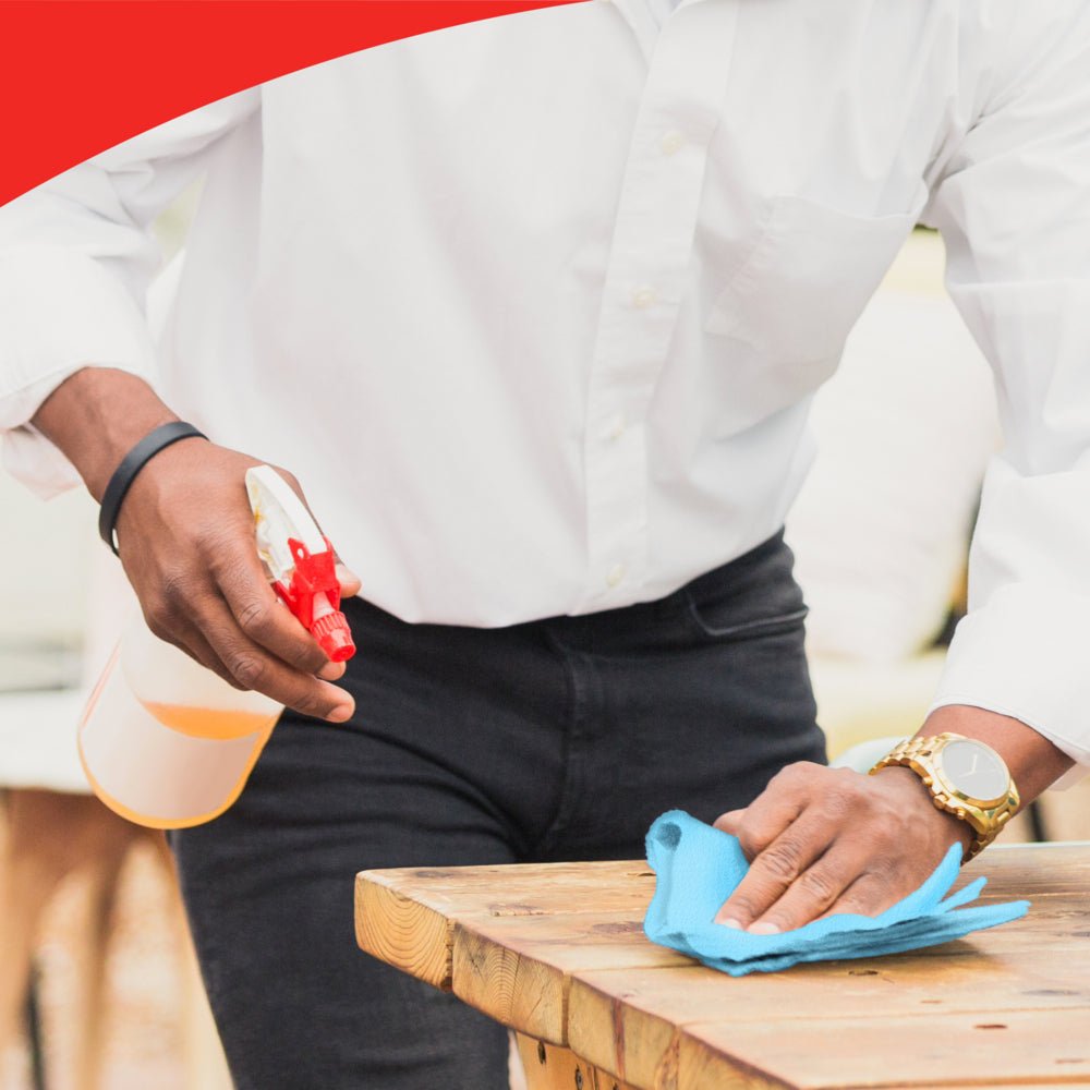 Osoba w białej koszuli i złotym zegarku czyści drewniany stół niebieską rolką WypAll® X60 General Clean od Kimberly-Clark GmbH. Jej twarz jest niewidoczna. Obraz ma czerwony pasek na górnym brzegu.
