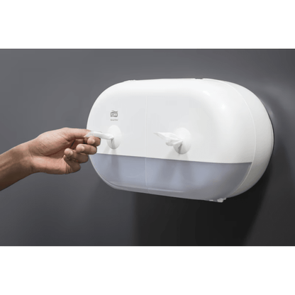 Osoba wyciąga papierowy ręcznik z eleganckiego białego Tork SmartOne® 682000 Mini podwójnego dozownika rolek papieru toaletowego, który posiada dwie komory i jest zamontowany na gładkiej, ciemnoszarej ścianie.