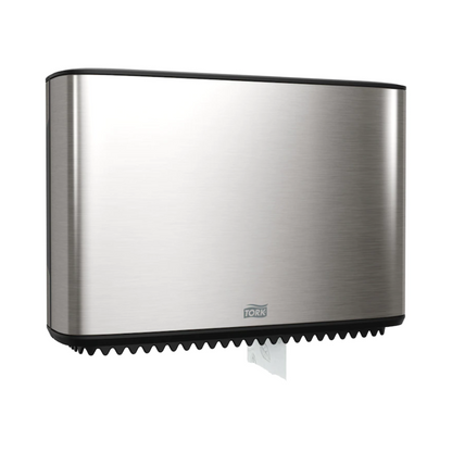 Dozownik Tork Xpress na papier toaletowy Mini-Jumbo, będący częścią Image Line, jest elegancko zamontowany na ścianie. Ten dozownik ze stali nierdzewnej ma smukły prostokątny design, z którego dolnej części widoczny jest mały fragment białego papieru. Marka „TORK – Essity Professional Hygiene Germany GmbH” jest wyraźnie widoczna na przedniej stronie.