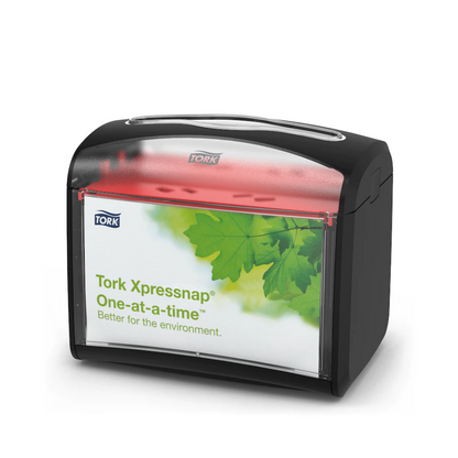 Dozownik na serwetki Tork Xpressnap® 272611 od TORK – Essity Professional Hygiene Germany GmbH ma czarną górę i przezroczysty przód z zielonym wzorem liści i napisem „Tork Xpressnap One-at-a-time – lepsze dla środowiska”. Ten dozownik poprawia higienę, jednocześnie efektywnie redukując zużycie serwetek.