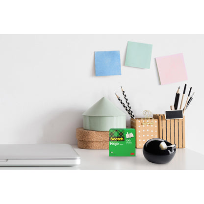 Biurko z zamkniętym laptopem, 3M Scotch™ Magic Tape, czarny Scotch™ Karim Rashid Tischabroller od 3M Deutschland GmbH, kubek z długopisami i ołówkami, stos okrągłych pudełek i trzy pastelowe karteczki samoprzylepne na białej ścianie.