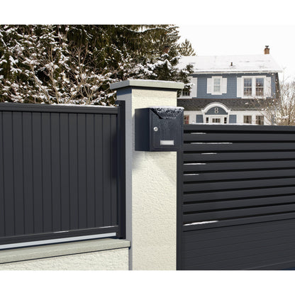 Ein moderner schwarzer Briefkasten mit Schnee ist auf einer weißen Säule neben einem Metallzaun montiert, befestigt mit Scotch-FIX™ Extreme doppelseitiges Außenmontageband von 3M Deutschland GmbH, mit einem blaugrauen Haus und verschneiten Bäumen im Hintergrund.