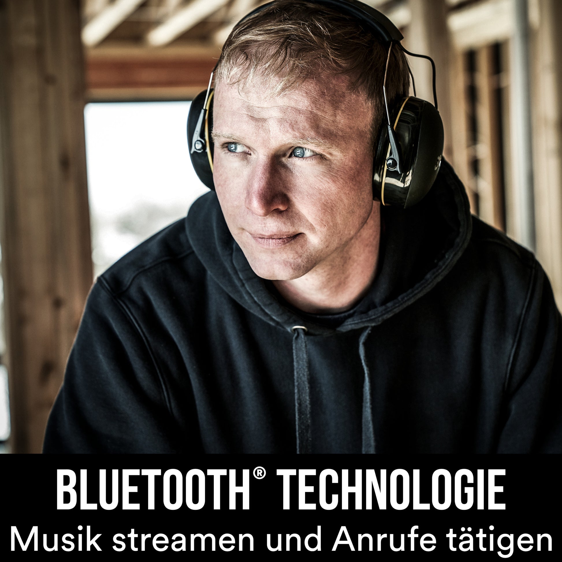 Ein Mann in einem schwarzen Kapuzenpulli trägt 3M™ WorkTunes™ Connect Kapselgehörschutz 90543E (SNR 31 dB, Bluetooth®, Schwarz) in einem Holzgebäude. Text: "Bluetooth Technologie. Musik streamen und Anrufe tätigen.