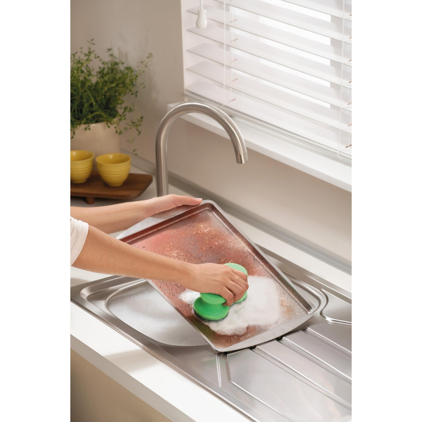 Ktoś szoruje duży, zabrudzony blachę do pieczenia za pomocą Scotch-Brite® Classic gąbki do czyszczenia 3M Deutschland GmbH pod kranem w jasnej kuchni z białymi żaluzjami, rośliną i ułożonymi żółtymi miskami w tle.