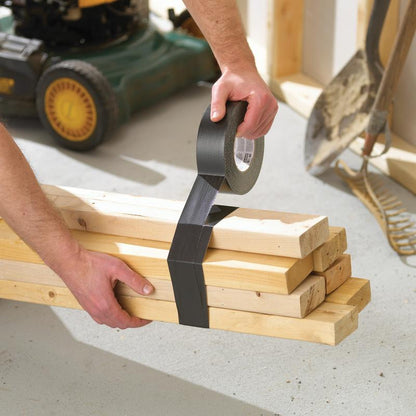 W warsztacie ktoś zabezpiecza drewniane deski za pomocą taśmy klejącej Scotch® Extremium™ Ultra Wysokiej Wydajności DT17 (10 m x 24 mm) od 3M Deutschland GmbH. W pobliżu stoją łopata, grabie i kosiarka.