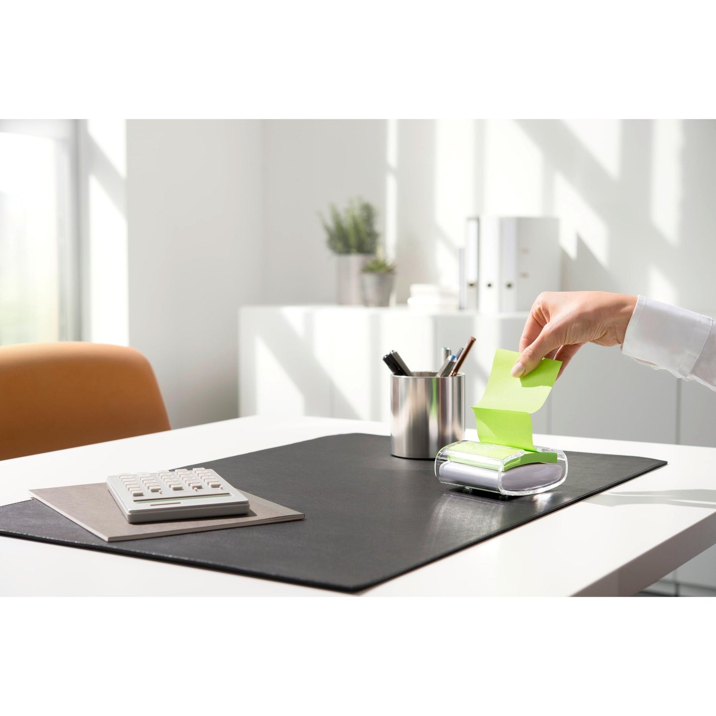 Osoba wyciąga żółtą karteczkę Post-it® Super Sticky Z-Note (76 x 76 mm) z czarnego podajnika PRO Post-it® Z-Notes firmy 3M Deutschland GmbH, na uporządkowanym biurku z kalkulatorem, pojemnikiem na długopisy i czarną podkładką w jasnym świetle dziennym.