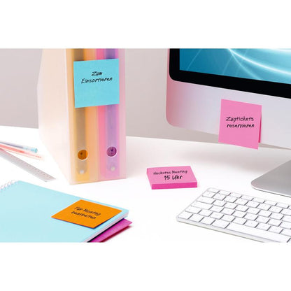 Scena biurowa z pastelowymi segregatorami, notatnikami i żółtymi karteczkami 3M Post-it® (76x76 mm, 90 arkuszy/blok, 6 bloków, 100% PEFC), które są przymocowane do materiałów biurowych i monitora, obok białej klawiatury i niebieskiego ekranu.
