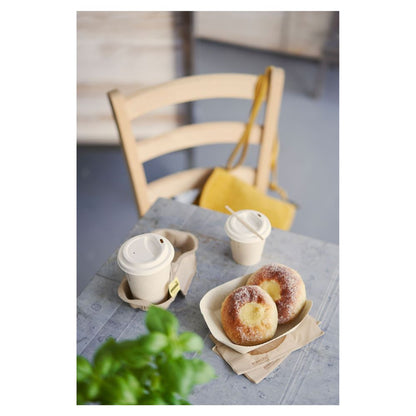 Na stole leżą dwa posypane cukrem ciastka na tacy i dwa kubki Duni „Sweet Bedruckte Bagasse” z pokrywkami firmy Duni GmbH. Żółta torebka wisi na drewnianym krześle, podczas gdy częściowo widoczna zielona roślina na pierwszym planie podkreśla subtelny temat zrównoważonego rozwoju.