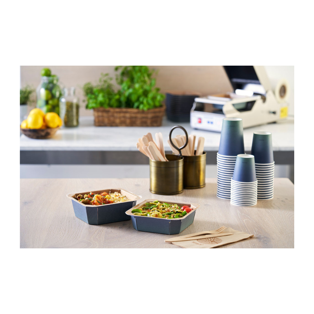 Na stole stoją dwie miski na wynos, otoczone ułożonymi kubkami do kawy Duni Urban PAP/PLA od Duni GmbH, drewnianymi sztućcami w uchwycie i serwetkami. Cytryny, szklanki i zioła w tle przyczyniają się do zrównoważonej atmosfery tego obszaru przygotowywania posiłków.